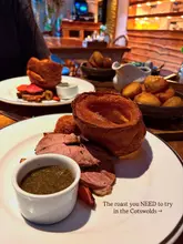 A close-up, slightly overhead shot of a Sunday roast featuring lamb, a large Yorkshire pudding, and roast potatoes, with a second plate visible in the background.
