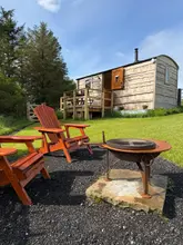 A bright, sunny outdoor shot of a wooden shepherd's hut with a fire pit and wooden chairs in the foreground, set in a grassy field.