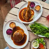 A high-quality, overhead flat-lay shot of a hearty meal featuring lamb shank, beef, mash, and broccoli. The image effectively captures the rustic, comforting nature of the food, making it look very appealing for a casual dining experience.