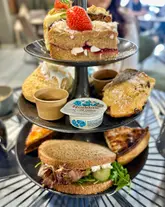 A close-up, eye-level shot of a three-tiered afternoon tea stand featuring a variety of sandwiches, pastries, scones, and cakes. The image is well-lit and clearly showcases the generous portion sizes, though the presentation is slightly marred by the inclusion of a plastic, branded clotted cream pot.