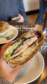 A short, handheld video review of Twelve Triangles in Edinburgh, showcasing their brunch offerings including a ham, cheese, and chutney sandwich, a yogurt and granola bowl, a cinnamon knot, and coffee.
