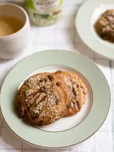 A high-quality, well-lit food photography shot featuring two homemade cookies topped with sesame seeds, presented on a simple plate with a beverage in the background. The image is part of a recipe tutorial for sugar-free, miso-infused oat flour cookies.