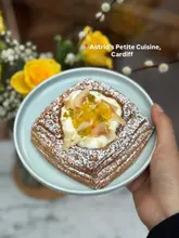 A high-quality, close-up shot of a mango, lime, and coconut Danish pastry held in a hand, showcasing the flaky texture and fresh toppings. The image uses soft, natural light and a shallow depth of field to make the pastry the clear, appetizing focus, effectively promoting the artisan nature of the pop-up venue.