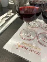 A close-up, slightly angled shot of a wine tasting mat at Manchester Wine School, featuring several glasses of red wine. The composition prioritizes the venue's logo on the mat over the wine itself.