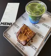A top-down, well-lit photograph of a blue matcha latte and a Biscoff brownie served on a branded tray at a cafe, with a menu visible in the background.