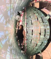A wide-angle, fisheye-style photograph taken inside an immersive art exhibition, featuring a person standing in the center of a mirrored, screen-covered room.