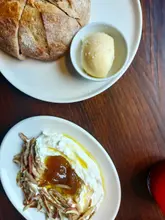 A top-down, overhead shot of a meal consisting of a loaf of bread with a side of butter and a bowl of feta whip topped with fig jam and pecans, set on a dark wooden table.