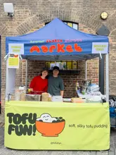 A friendly, approachable photo of two vendors standing behind their 'Tofu Fun' market stall, effectively introducing their brand and presence at a street food market.