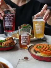 A high-quality, professional promotional photograph featuring two people holding branded glasses from Makars Gourmet Mash Bar Company. In the foreground and background, portions of their signature mash-topped pie are visible, creating an inviting and cohesive brand image.