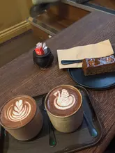 A high-angle shot of a cafe table featuring two mugs of hot chocolate with latte art and a slice of banana bread on a black plate, accompanied by a small cauldron-shaped sugar holder.