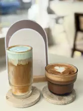 A high-quality, well-lit shot of two specialty drinks—a layered iced latte with a blue-tinted foam and a hot mocha with latte art—placed on stone coasters in a cafe setting.