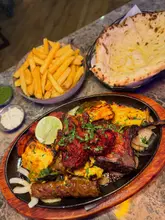 A top-down shot of a large Indian sizzler platter featuring lamb chops, prawns, chicken, and paneer, accompanied by a bowl of masala chips and a cheesy garlic naan.