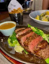 A close-up shot of a sliced fillet steak served with greens and a side of peppercorn sauce, accompanied by a cone of fries and a bowl of pasta in the background.