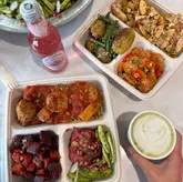 A bright, overhead flat-lay photograph showcasing a healthy takeaway lunch spread. The image features several containers filled with Mediterranean and Middle Eastern-inspired dishes, including chicken meatballs and various salads, accompanied by a bottle of rose lemonade and a matcha latte held by the creator.