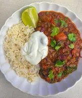 A top-down, flat-lay photograph of a bowl of homemade chilli con carne served with rice, a dollop of sour cream, and a lime wedge, garnished with fresh coriander.