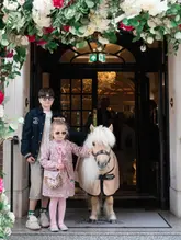 A high-quality, professional lifestyle photograph taken at the entrance of The Goring hotel, featuring two children posing with a Shetland pony under a floral archway. The image conveys a sense of luxury, family-friendly hospitality, and whimsical charm.