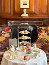 A beautifully composed, symmetrical shot of a luxury afternoon tea service set against a classic, wood-paneled fireplace. The image captures the elegance of the venue, with the tea stand, champagne flutes, and delicate pastries arranged perfectly to evoke a sense of high-end hospitality.