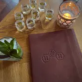 A high-quality, warm-toned flat lay photograph of a table setting at Hive & Honey, featuring a leather-bound menu, a cocktail, and several small glasses containing what appear to be shots or tasters, illuminated by a candle.