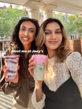 A bright, casual selfie-style photo of two friends holding colourful drinks outside a cafe, promoting a relaxed social atmosphere.