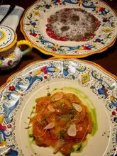 A high-quality, overhead shot of two starters at Patatino in Edinburgh, featuring a beef carpaccio with truffle and a salmon crudo, served on ornate, colorful ceramic plates on a dark wooden table.