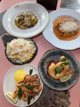 A high-quality, overhead flat-lay photograph of a spread of Italian dishes, including pasta, lasagne, calamari, king prawns, and parmesan fries, arranged on a pink table.