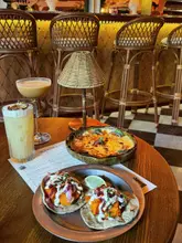 A high-quality, atmospheric shot of a table spread at a restaurant, featuring two cocktails, a small bowl of dip, and a plate of two tacos. The lighting is warm and inviting, perfectly capturing the aesthetic of the venue.
