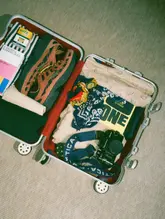A flat-lay photograph of an open suitcase packed with travel essentials, including sneakers, a camera, a book, and clothing, intended to promote a new aparthotel.