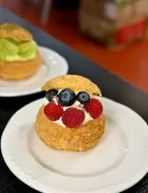 A close-up shot of a cream puff topped with fresh blueberries and raspberries, served on a white plate in a casual market setting.
