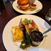 A close-up shot of a duo of pork dish featuring pork belly and fillet with dauphinoise potatoes and a side of gravy, with a chicken burger visible in the background at a restaurant.