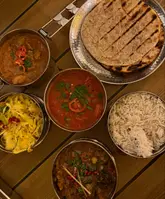 A top-down, overhead shot of a 'Street Food Sharer' meal from Mowgli, featuring various curries, rice, roti, and sides served in traditional metal tiffin tins on a wooden table.