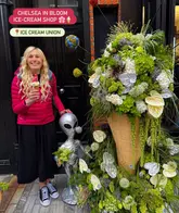 A photo of a person holding an ice cream cone while standing next to a large floral display (Chelsea in Bloom) and an alien statue outside an ice cream shop.