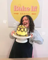 A bright, promotional photo featuring a creator holding a bee-themed cake at a baking experience venue in Bristol, effectively showcasing the fun and interactive nature of the activity.