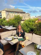 A lifestyle shot featuring a woman enjoying a glass of white wine at a rooftop bar in Soho, London. The setting is an outdoor terrace with greenery and city views, capturing a relaxed, upscale atmosphere.