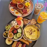 A high-angle flat lay shot of a generous brunch spread featuring two large plates filled with a variety of breakfast items, including eggs, sausages, halloumi, hummus, and fruit, accompanied by iced drinks.