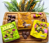 A clean, well-lit flat-lay photograph showcasing M&S chocolate products, including 'The Big Daddy' Pistachio Bites, Caramel Filled Buttons, and Speckled Eggs, arranged on a wooden board with decorative dried flowers.
