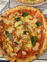 A top-down shot of a Neapolitan-style pizza, likely a Margherita, featuring melted cheese, tomato sauce, and fresh basil leaves. The pizza is served on a table with a menu visible underneath, capturing a casual dining experience.