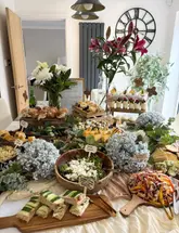 A high-angle shot of a beautifully arranged grazing table featuring a variety of finger foods, including sandwiches, quiche, salads, and desserts, set up in a domestic dining area.