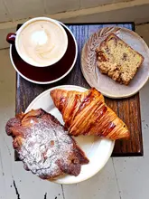 A high-quality, overhead flat-lay photograph of a cafe spread featuring a cup of coffee, a slice of honey walnut cake, a plain croissant, and a chocolate almond croissant arranged on a rustic wooden table.