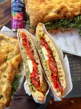 A high-quality, appetizing close-up shot of a freshly made Italian panuozzo sandwich, held up to highlight the texture of the bread and the generous fillings. The lighting is natural and flattering, effectively showcasing the food's quality, while the background provides relevant context without distracting from the main subject.