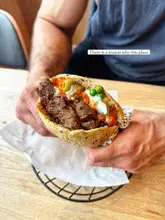 A high-quality, close-up shot of a filled pita sandwich held by a person, showcasing the textures of the meat, sauce, and fresh toppings. The lighting is bright and natural, highlighting the food's appeal against a simple, clean background.