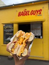 A close-up, handheld shot of a bacon, egg, and cheese bagel held in front of a bright yellow 'The Bagel Guys' market stall. The focus is sharp on the textures of the bagel and the melted cheese, with the background softly blurred to keep attention on the food.