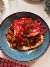 A high-quality, top-down photograph of a breakfast hash served in a blue ceramic bowl, featuring crispy potatoes, a poached egg, pickled red onions, and a creamy sauce base, set against a clean white table.