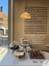 A bright, aesthetic shot of a cafe counter featuring a selection of cakes under glass domes and a menu board on the wall. The image is styled to appeal to a 'wellness' demographic, emphasizing healthy, refined sugar-free options.