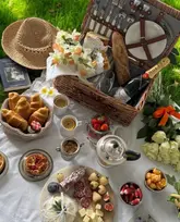 A high-quality, aesthetically pleasing flat-lay photograph of a luxury picnic spread, featuring a wicker hamper, champagne, croissants, strawberries, charcuterie, and fresh flowers on a white tablecloth set on grass.