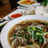 A close-up, 45-degree angle shot of a bowl of beef brisket pho, with other Vietnamese dishes like chicken curry and prawn crackers visible in the blurred background.