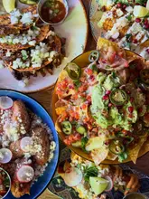 A vibrant, overhead flatlay shot of a Mexican-inspired vegan feast, featuring a spread of tacos, nachos, corn, and other small plates. The lighting is bright and natural, highlighting the fresh ingredients and textures of the food.