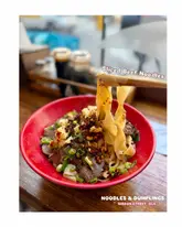 A close-up, eye-level shot of a bowl of sliced beef noodles being lifted with chopsticks, set against a wooden table in a restaurant. The image captures the texture of the noodles and the toppings, though the background is cluttered with condiment bottles.