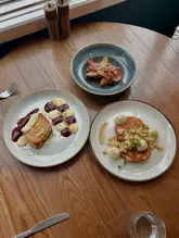 A top-down, overhead shot of a brunch spread featuring three distinct plates: eggy bread with bacon, buttermilk pancakes with fruit, and a 'Manchester Toast' dessert dish, set on a wooden table.