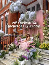 A high-quality, well-composed lifestyle photograph featuring a creator posing in front of an elaborate floral installation for the Chelsea in Bloom event. The image is bright, sharp, and effectively captures the aesthetic appeal of the event, serving as an invitation for viewers to visit the location for photo opportunities.