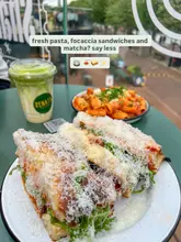 A top-down, slightly angled shot of a lunch spread featuring a chicken parm focaccia sandwich, a bowl of rigatoni, and a matcha latte, set on a green table in a food hall environment.